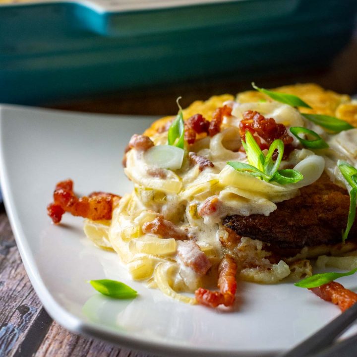 A slice of Kugelis on a white square plate with the sour cream onion and bacon sauce on top.