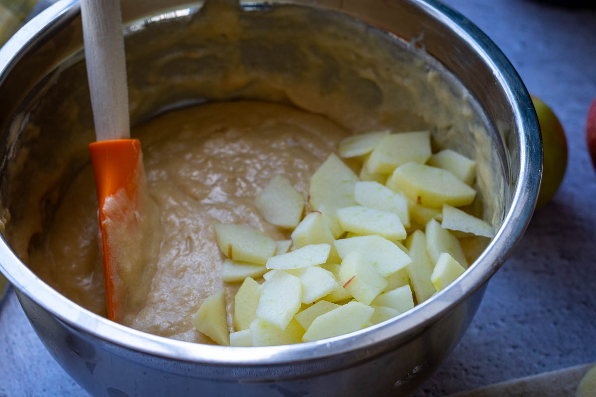 Thin apple slices being mixed into the cake batter in a metal bowl.