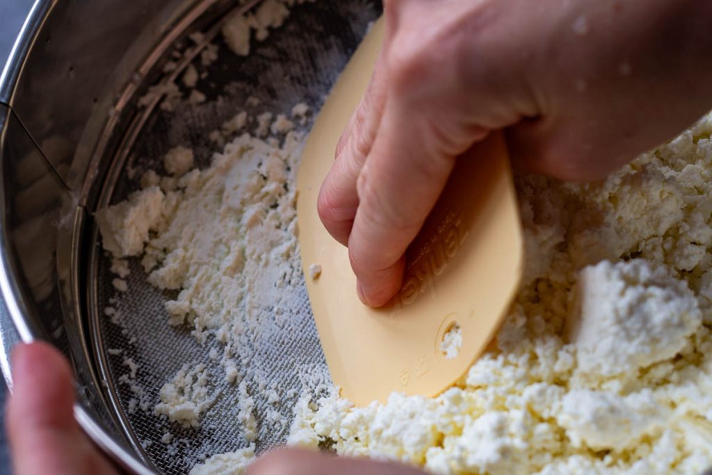 Pushing farmer's cheese through a sieve using a flexible dough scraper. 