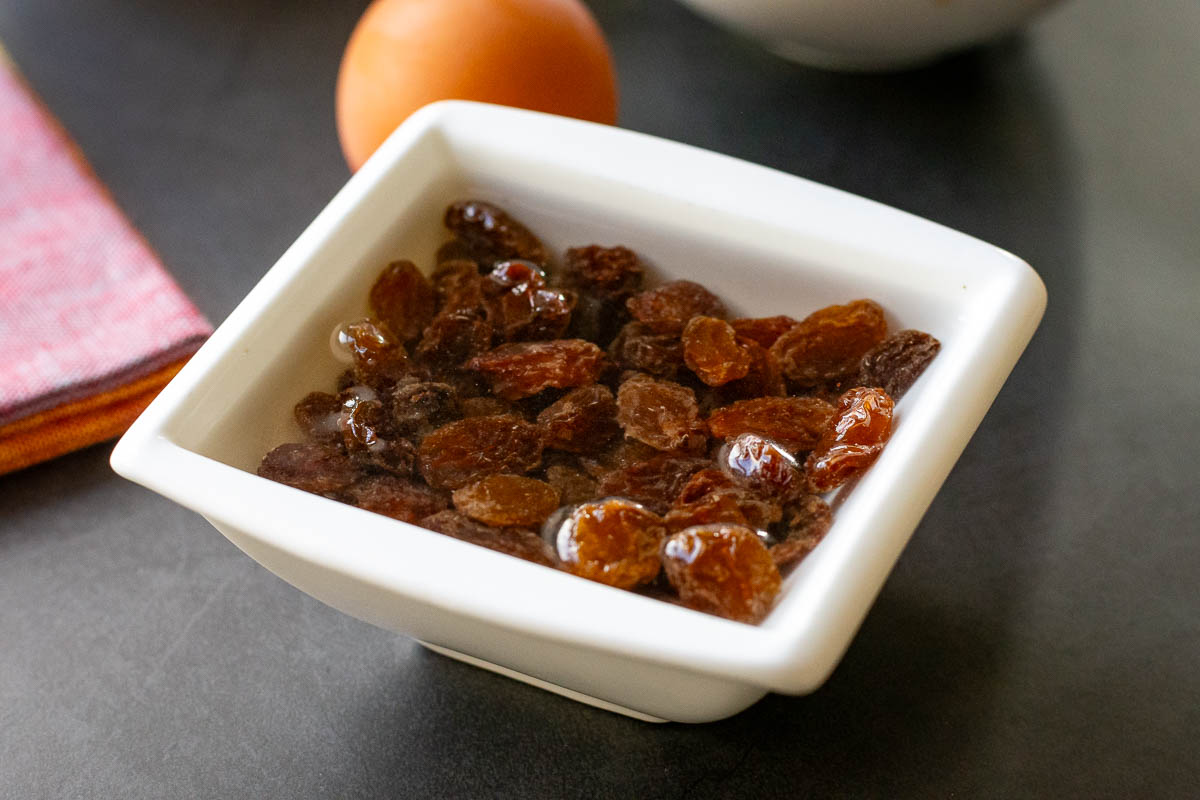 Sultanas soaking in hot water in a small square white bowl. 