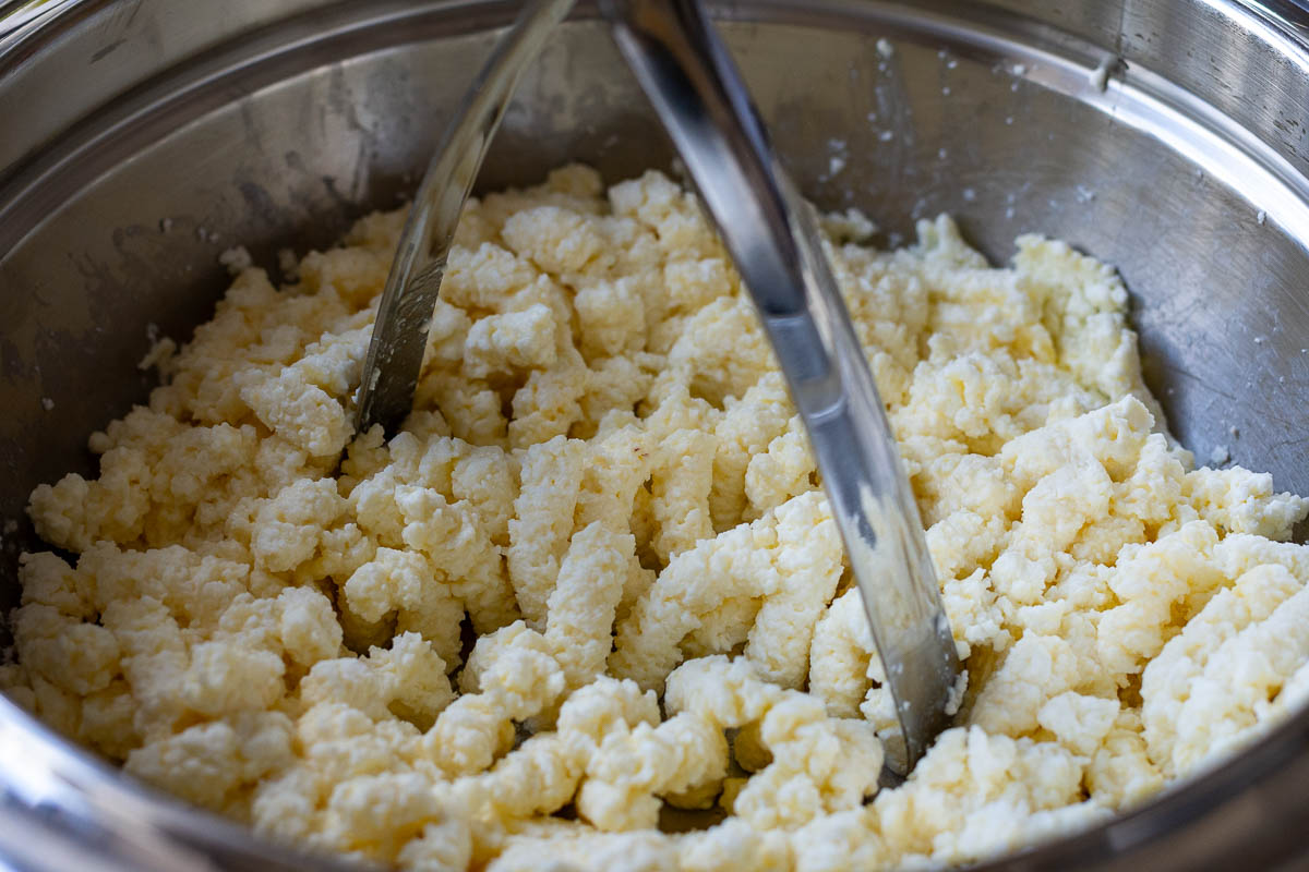 Mushing the wet ingredients for Lithuanian pancakes with a potato masher in a metal bowl. 