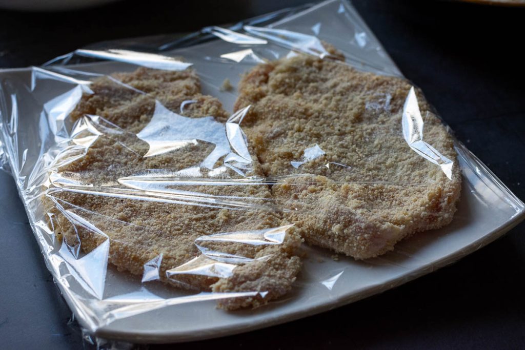 Two pork cutlets on a white plate covered with plastic wrap.
