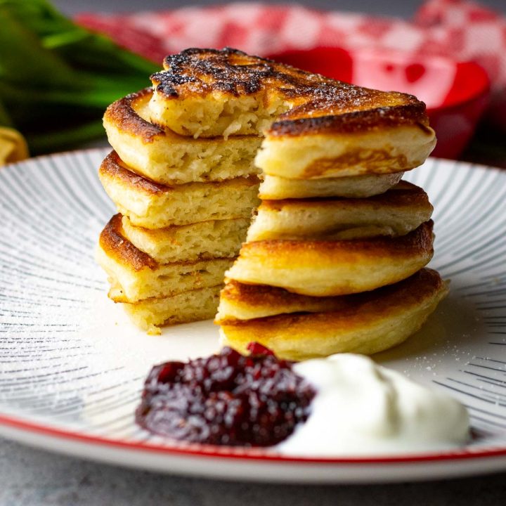A stack of 6 Lithuanian Kefir Pancakes with a wedge cut out on a white striped plate with red rim.