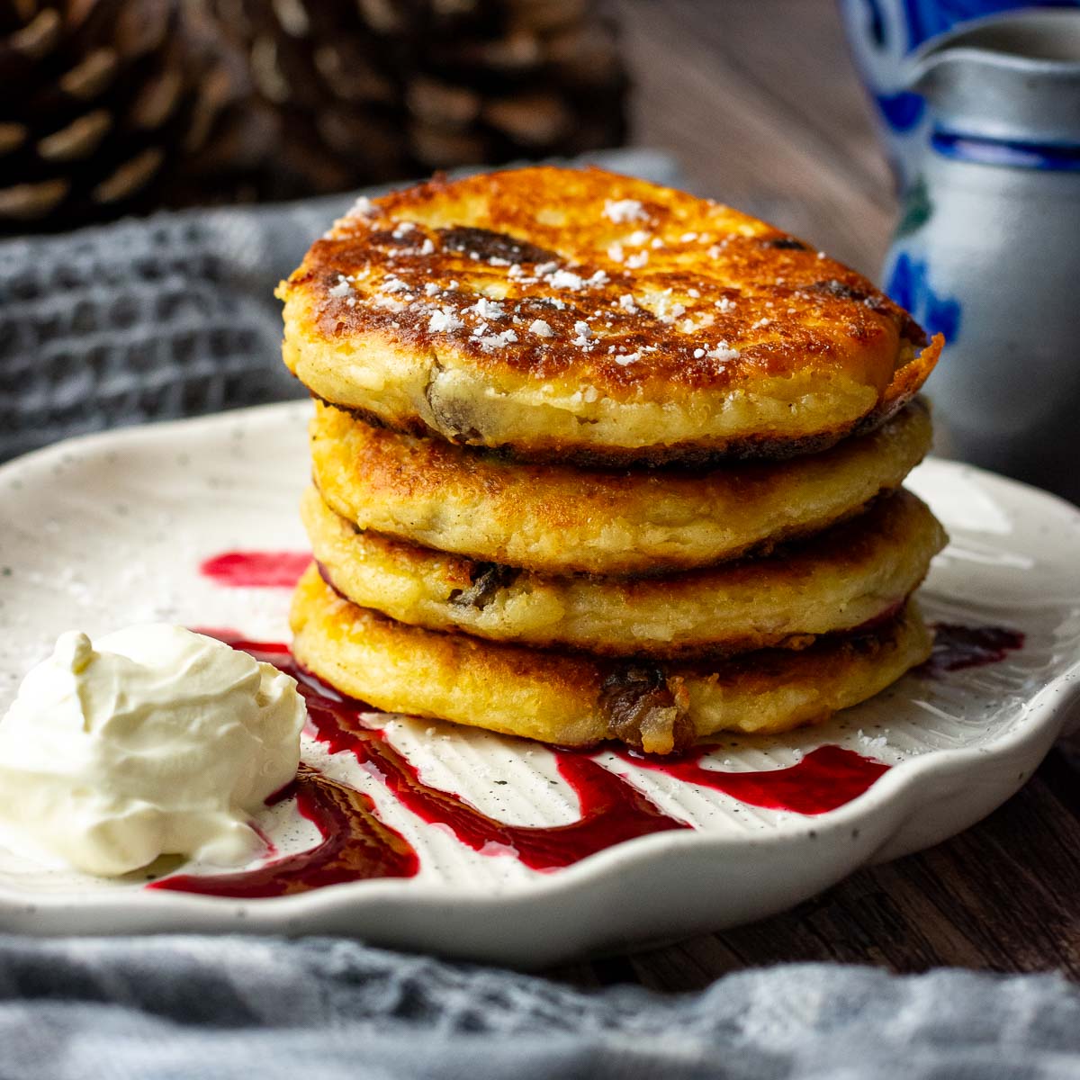 A stack of Lithuanian Farmer's Cheese Pancakes on a plate with sour cream and berry coulis.
