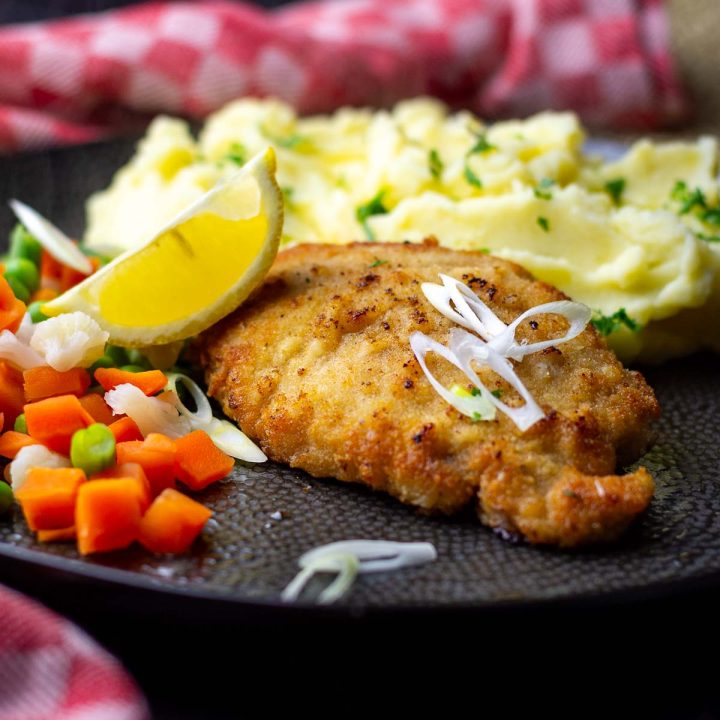 Lithuanian Pork Escalope on a black plate served with steamed vegetables, mashed potatoes and a wedge of lemon.