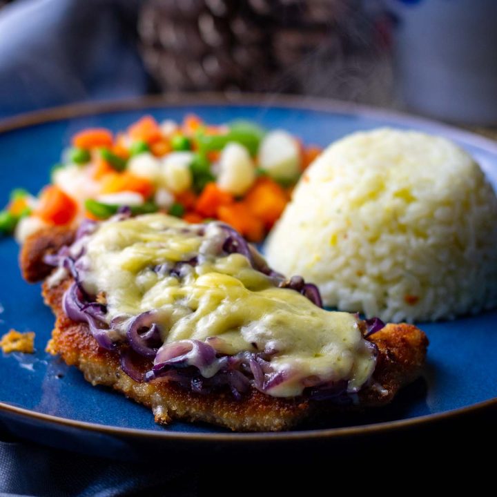 Breaded pork cutlet topped wth cheese and melted cheese served on a blue plate with rice and vegetables.