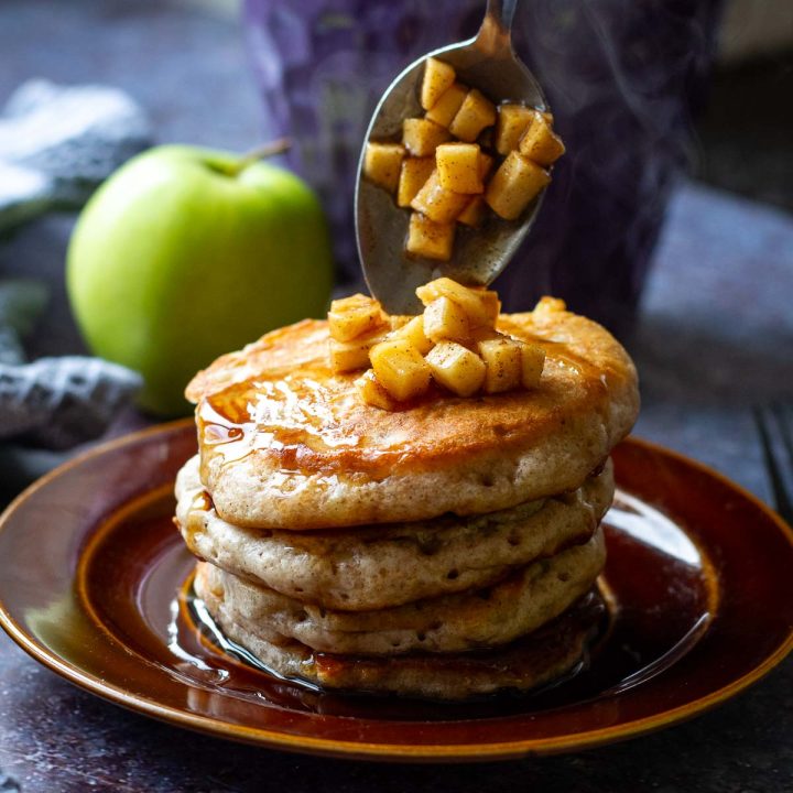 A stack of four grated apple pancakes being topped with cubes of stewed apples.