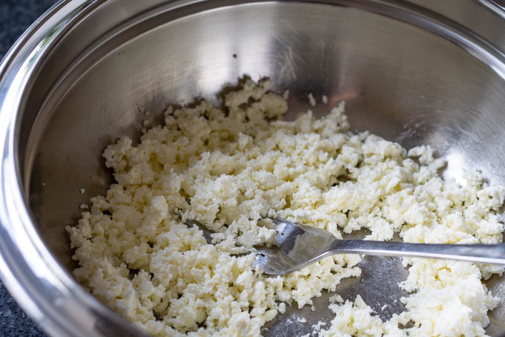 Mashing farmer's cheese with a fork in a metal bowl.