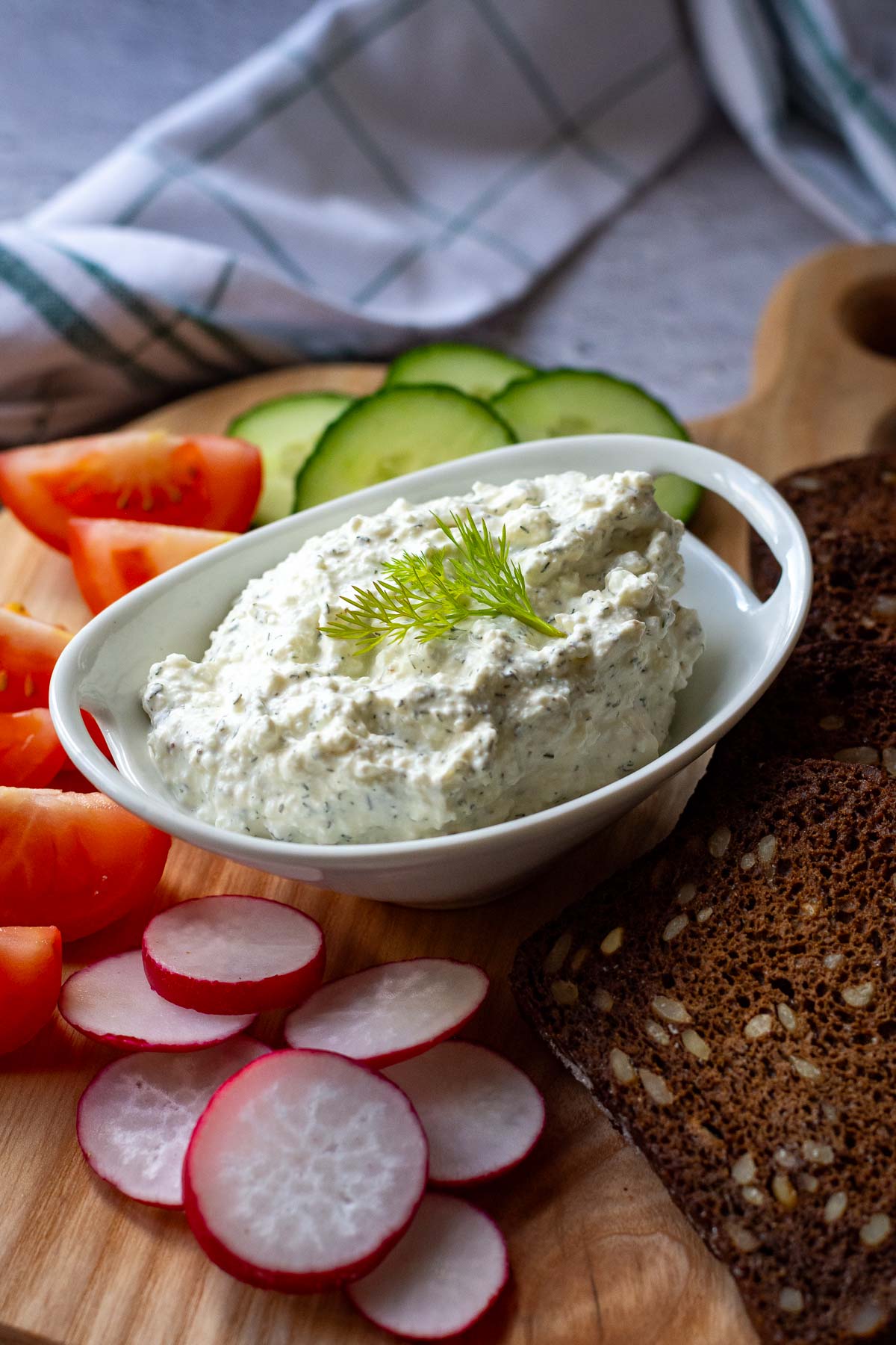 Dip with farmers cheese, sour cream and dill in a small white bowl amonds fresh veggies and rye bread slices.