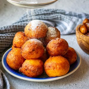 A pyramid of Lithuanian Farmer's Cheese Doughnuts on a small blue plate being dusted with powdered sugar.