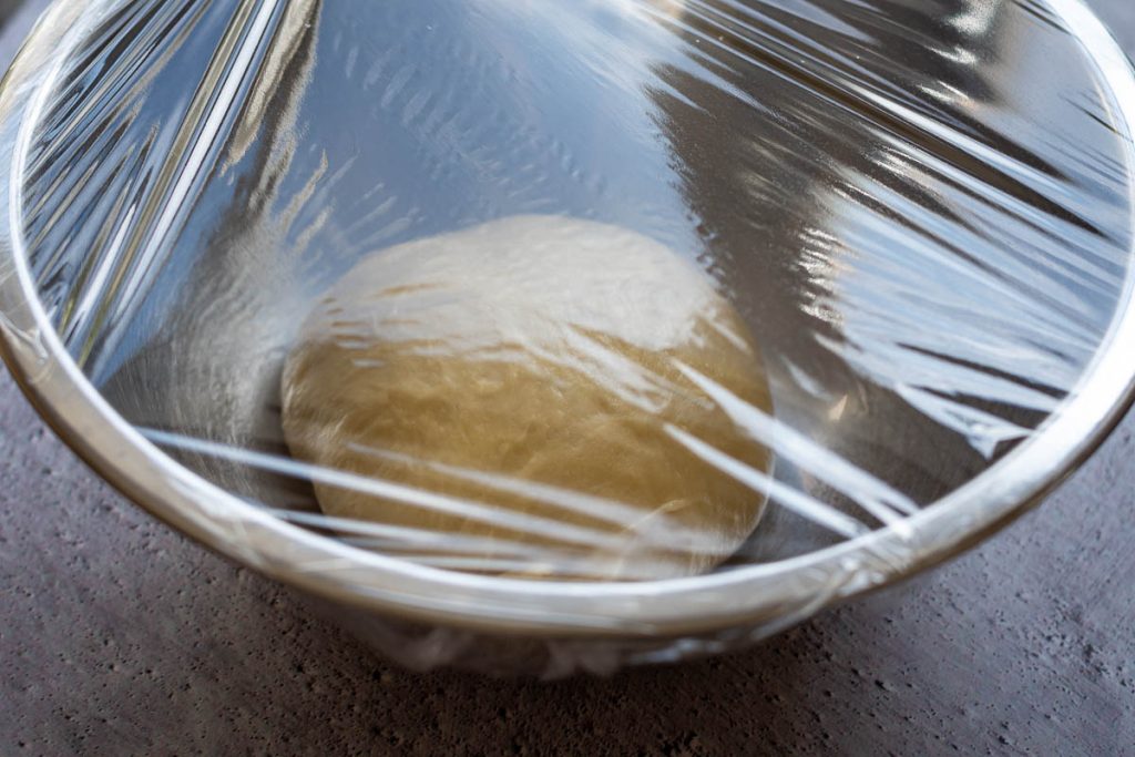 Lithuanian bun dough proofing in a large metal bowl covered with plastic wrap. 