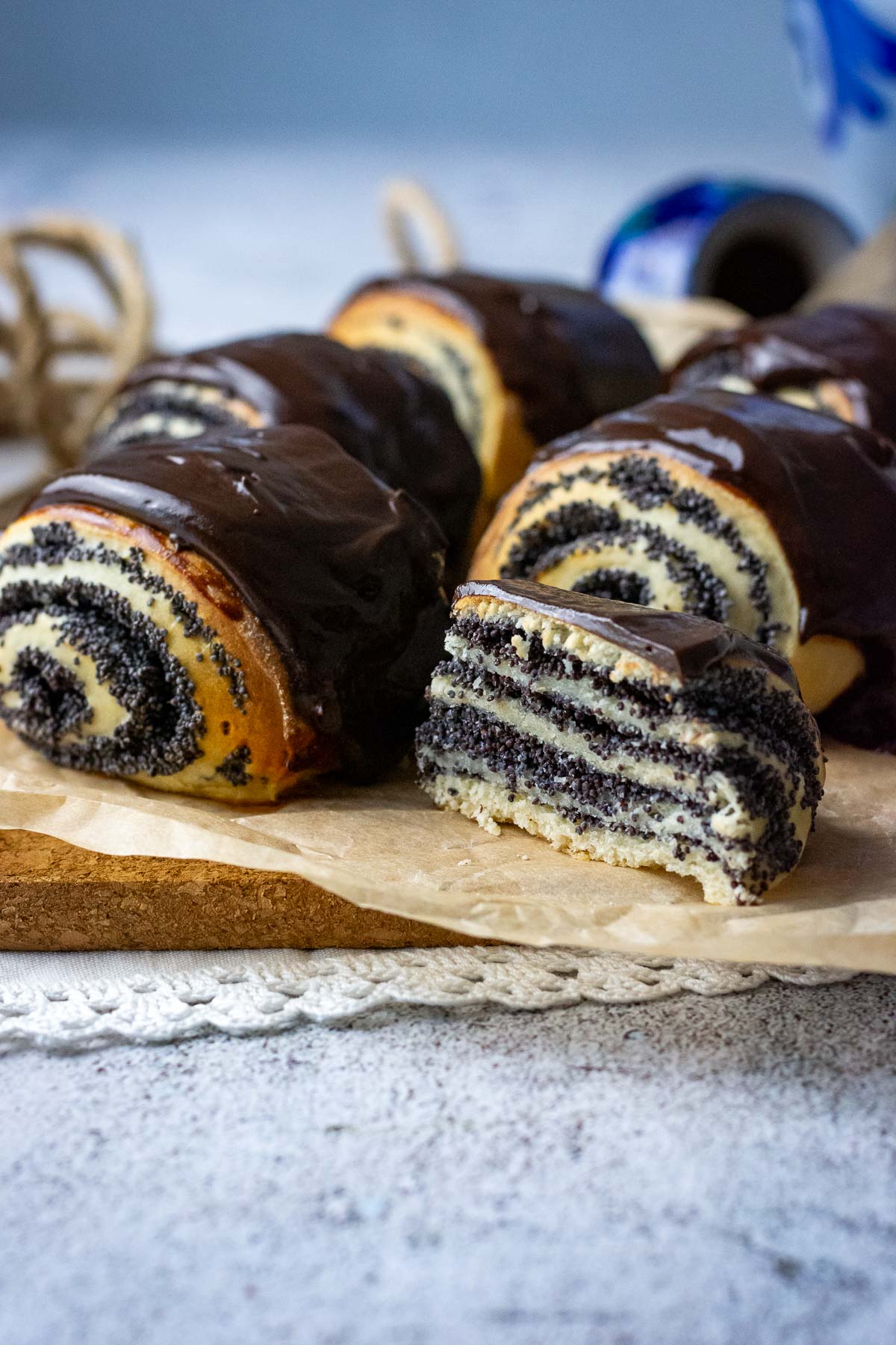 Chocolate glazed Lithuanian poppy seed rolls on a wooden board, one of them cut in half exposing the layers of poppy seed filling. 
