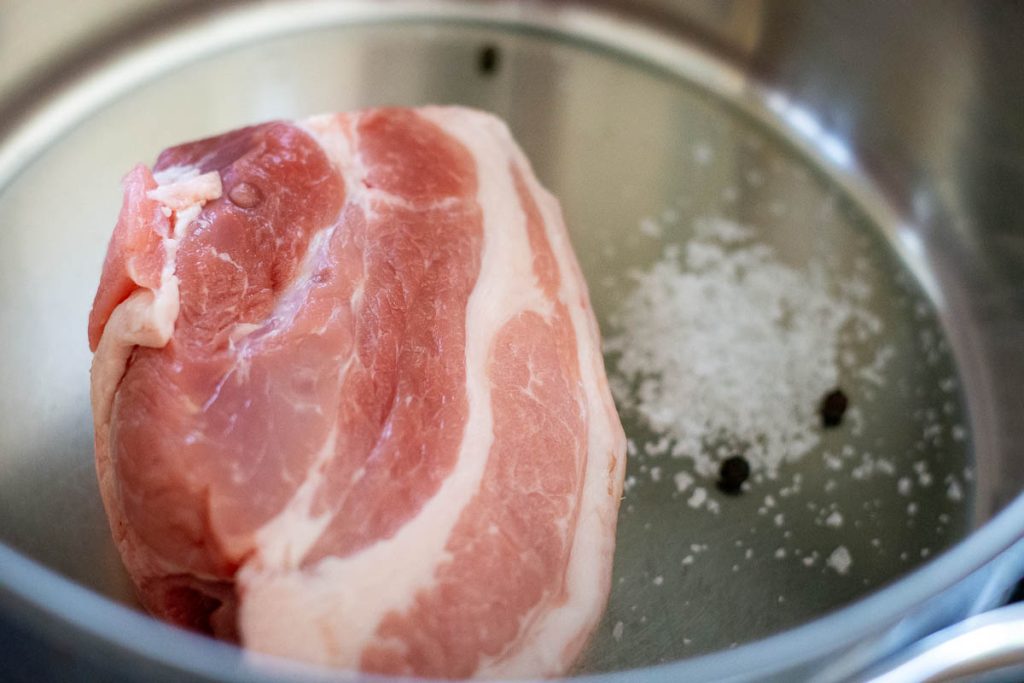 A piece of pork shoulder in a pan with salt a peppercorns ready to be boiled.
