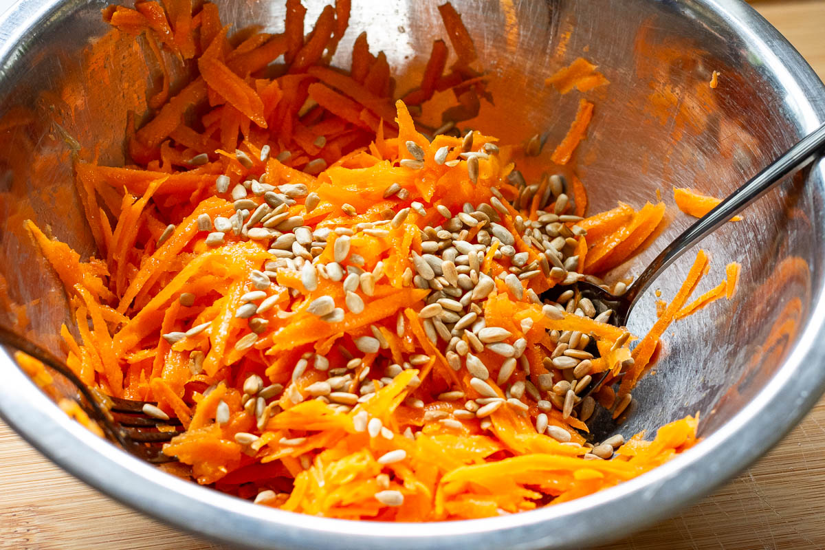 Grated carrots with toasted sunflower seeds on top in a metal bowl.