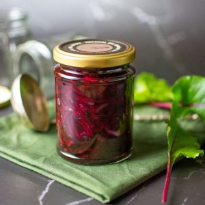 A jar of canned shredded beetroot on a green cloth surrounded by beetroot leaves.