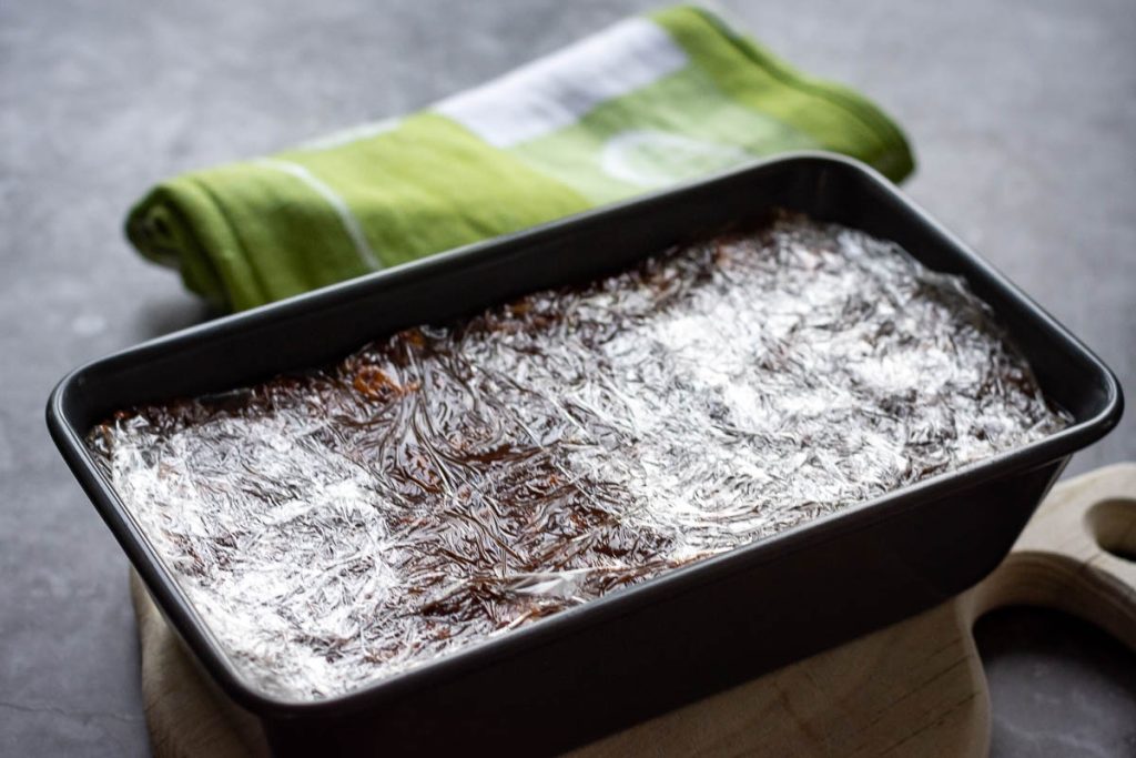 Lithuanian lazy cake in a loaf tin with the top covered with plastic wrap ready to be set in the fridge.
