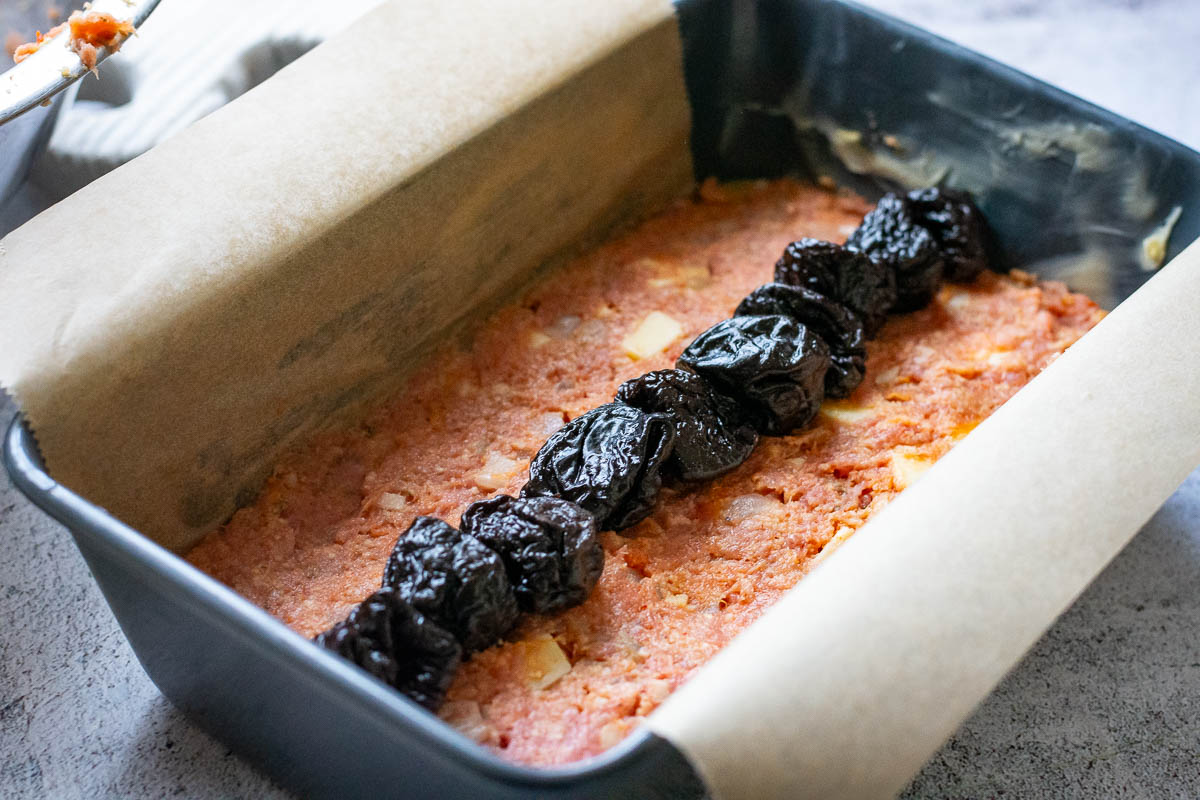 Prunes arranged on the first half of the meatloaf mixture pressed into a loaf pan.