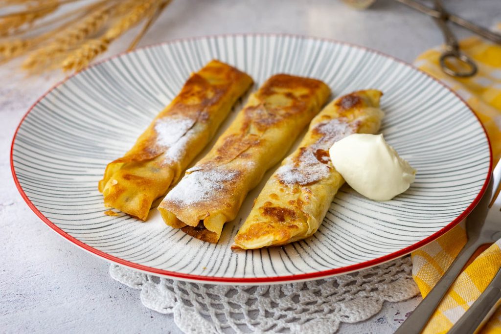 Three banana crepes with chocolate served with sour cream on a white striped plate.