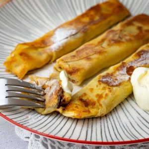 Three Banana crepes with chocolate on a white striped plate, showing melted chocolate and gooey banana inside the crepe.