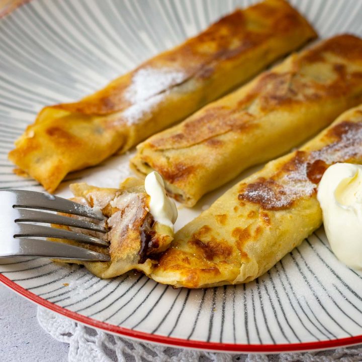 Three Banana crepes with chocolate on a white striped plate, showing melted chocolate and gooey banana inside the crepe.