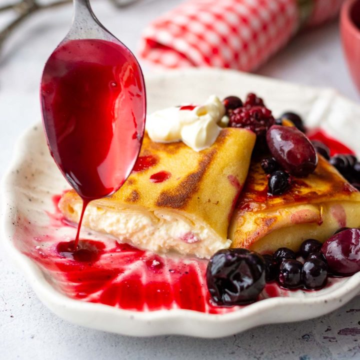 Two Lithuanian crepes with farmer's cheese filling being drizzled with berry compote.