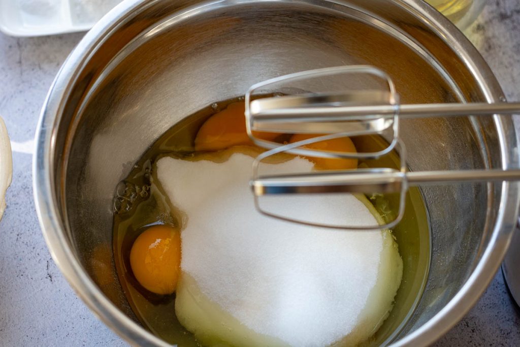Eggs and sugar in a mixig bowl with electrc hand mixer hovering above them. 