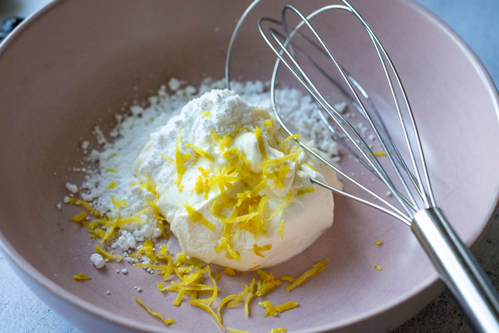 Creme fraiche, lemon zest and powdered sugar in a pale pink bowl with a balloon whisk in it. 