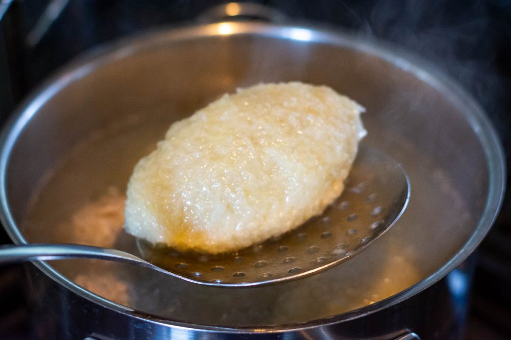 Cooked Lithuanian Cepelinas being lifted out of a pot of water with a slotted spoon.