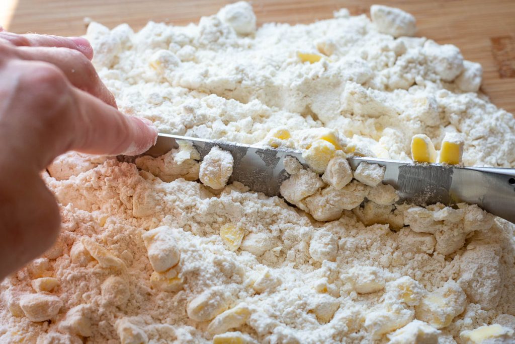 Cutting the cold butter into the flour mixture on a wooden board with a large knife.