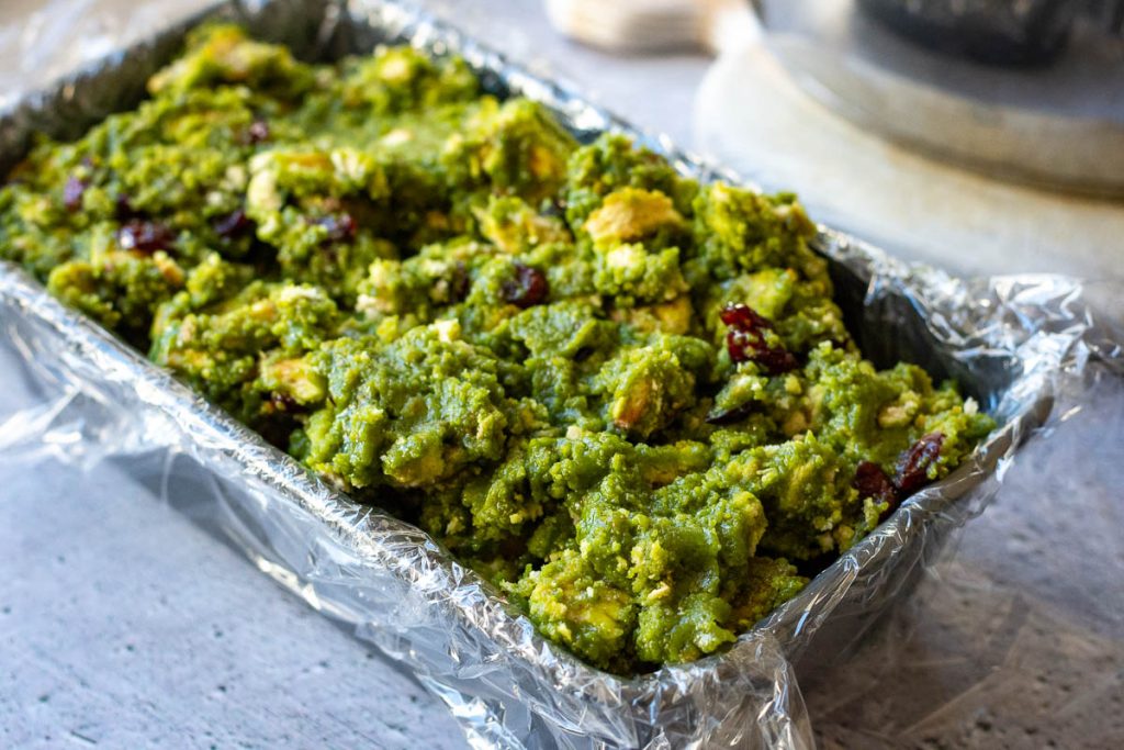 No bake matcha cake misture spooned into a loaf pan lined with plastis wrap.
