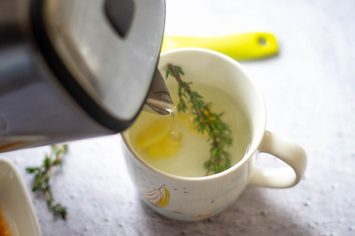 Pouring hot water from a kettle over the thyme and ginger. 