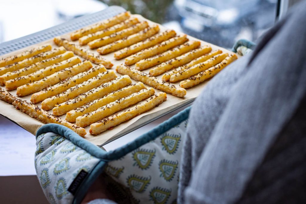 A babking sheet with cheese starws with caraway seeds held with oven gloves. 