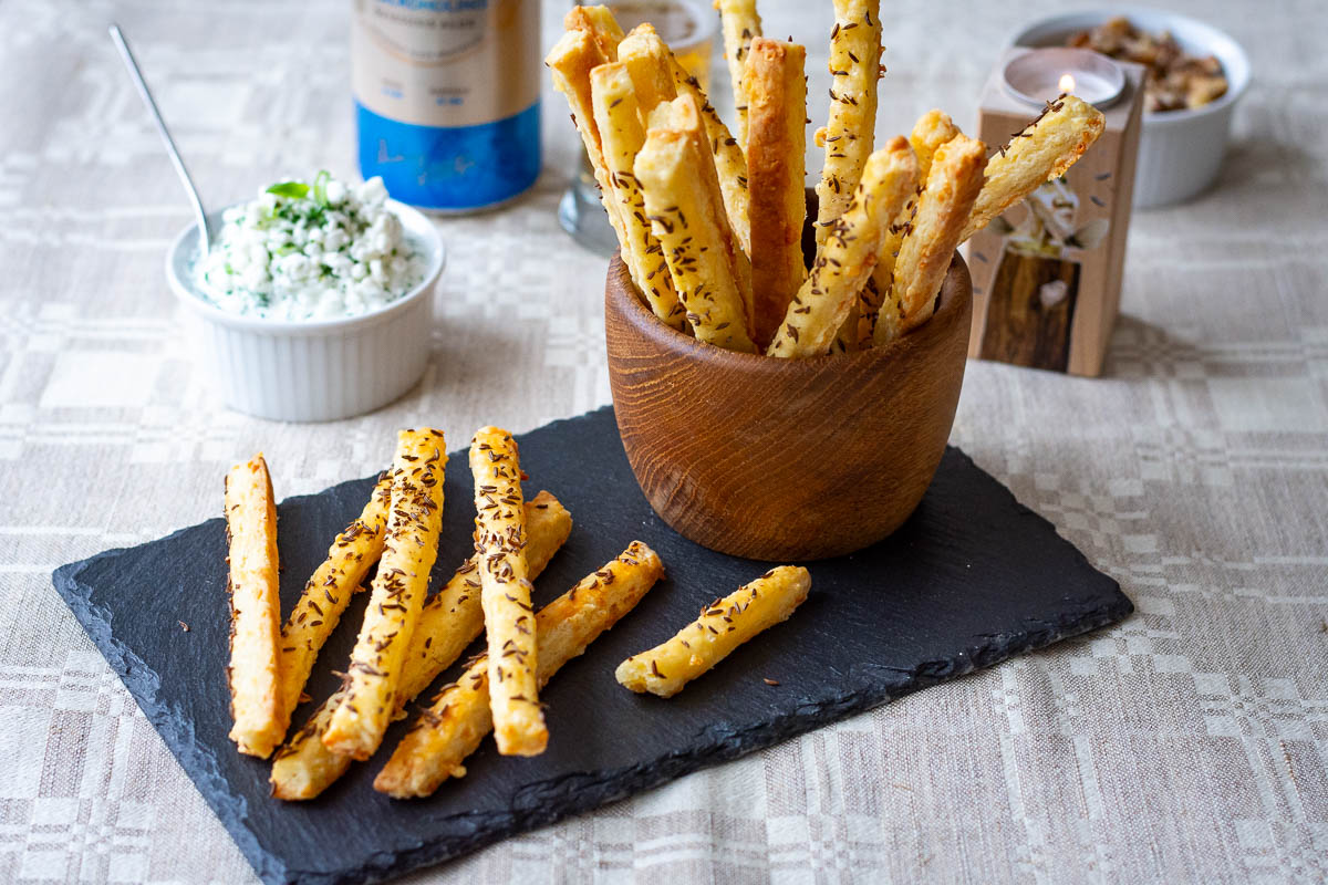 Lithuanian Cheese Straws with Caraway Seeds arranged on a black slate board with the rest of the cheese sticks in a wooden bowl. 