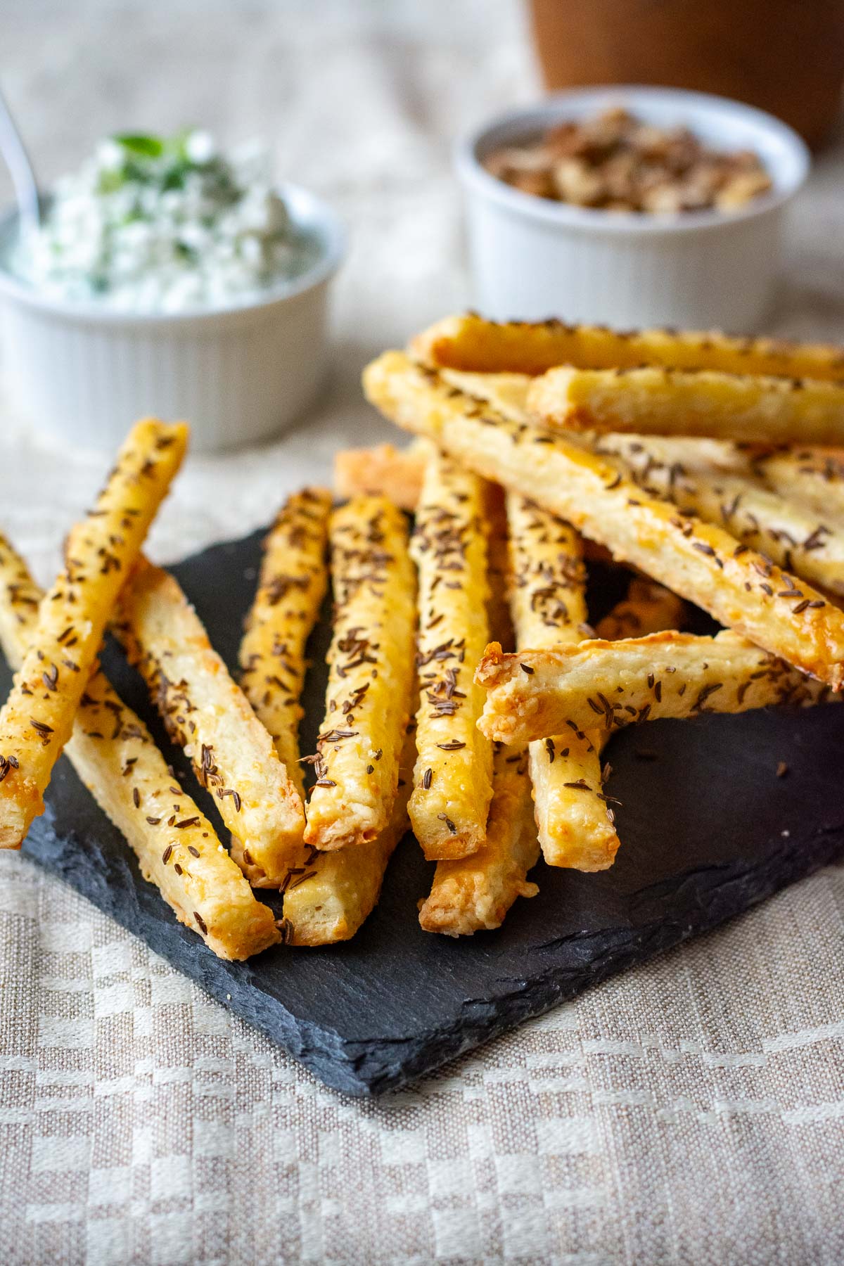 Lithuanian Cheese Straws with Caraway Seeds arranged on a black slate board with the rest of the cheese sticks in a wooden bowl.