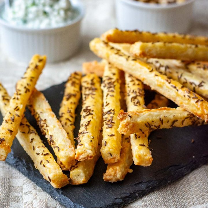 Lithuanian Cheese Straws with Caraway Seeds arranged on a black slate board with the rest of the cheese sticks in a wooden bowl.