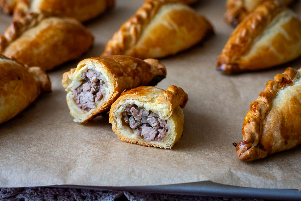 Lithuanian kibinai on a parchment paper lined baking sheet with one Lithuanian meat pie cut in half exposing savory pork filling.