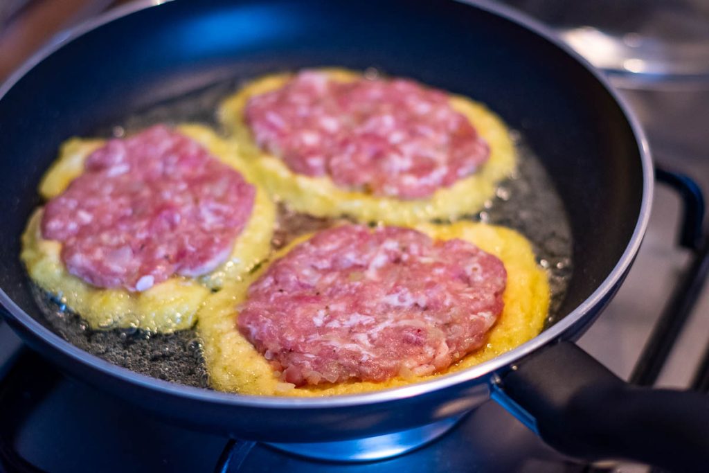 Grated potato pancakes topped with ground meat patty in a frying pan.